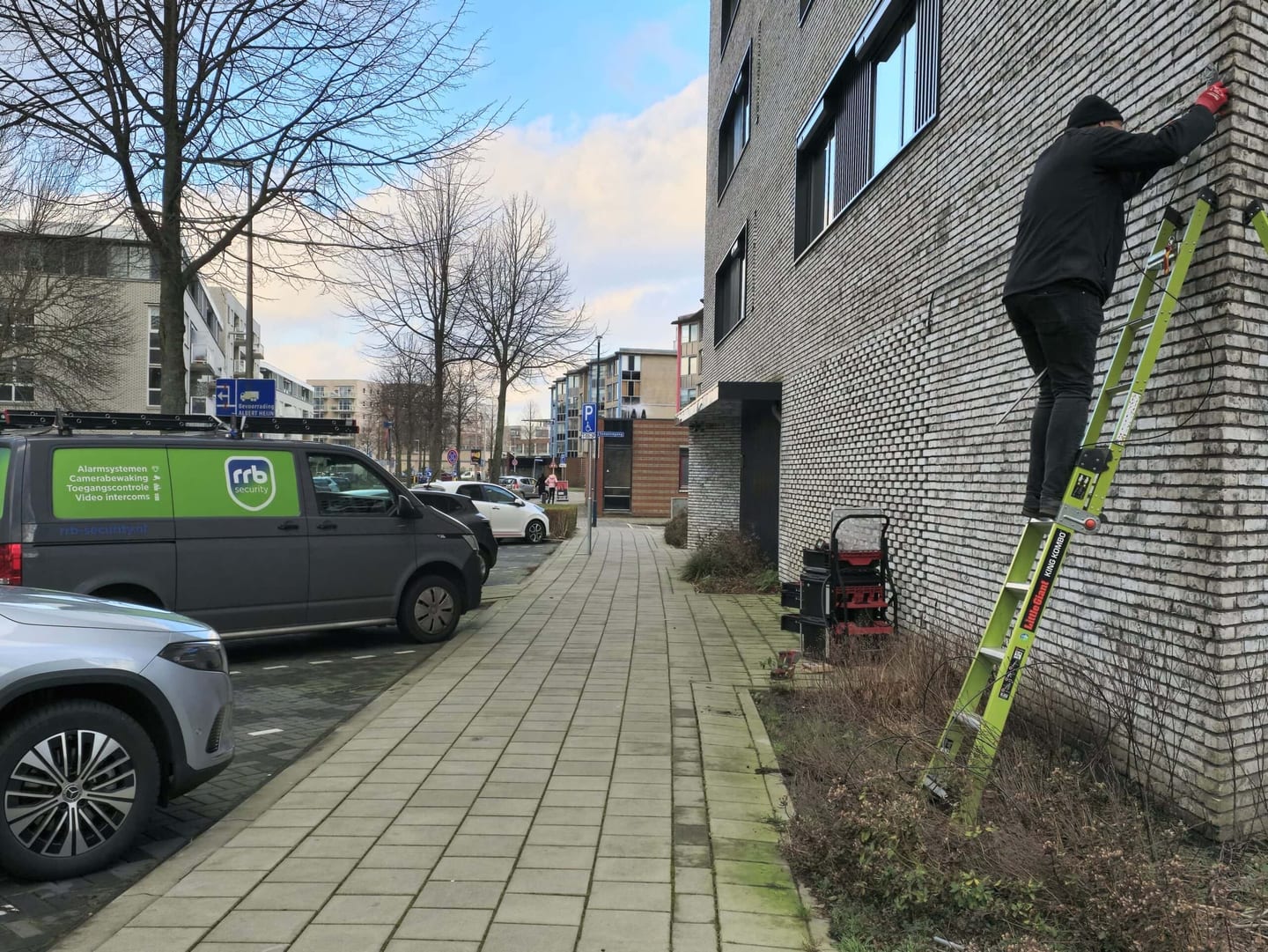 Contact RRB Security: monteur op een ladder tegen een bakstenen gevel, met bedrijfsbussen van RRB Security in de straat