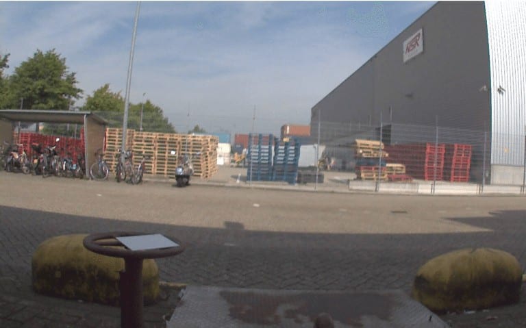 50+ Bewakingscamera Tips en Adviezen: camerabeeld van een bedrijfsterrein met pallets, fietsen en een grijze loods onder een blauwe lucht